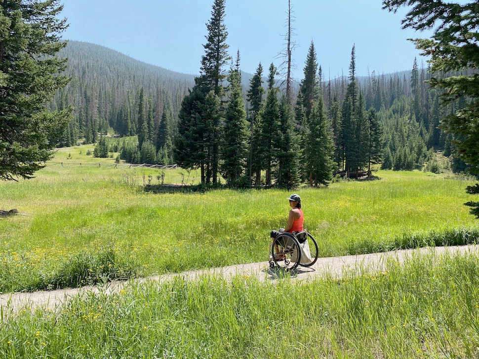 lady manual wheelchair user on trail through meadow with evergreen trees in the background